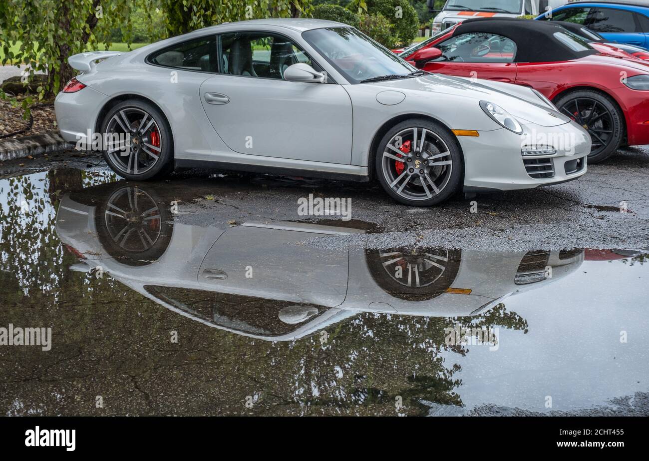 Nuova Porsche bianca in un parcheggio sotto un albero in un giorno piovoso con una riflessione speculare in a. grande puddle a terra all'aperto Foto Stock