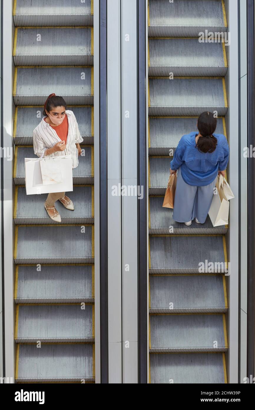 Vista dall'alto verso il basso di due donne che cavalcano la scala mobile in direzioni opposte mentre fanno shopping nel centro commerciale, concentratevi sulla donna che indossa la maschera guardando la fotocamera, spazio per la copia Foto Stock