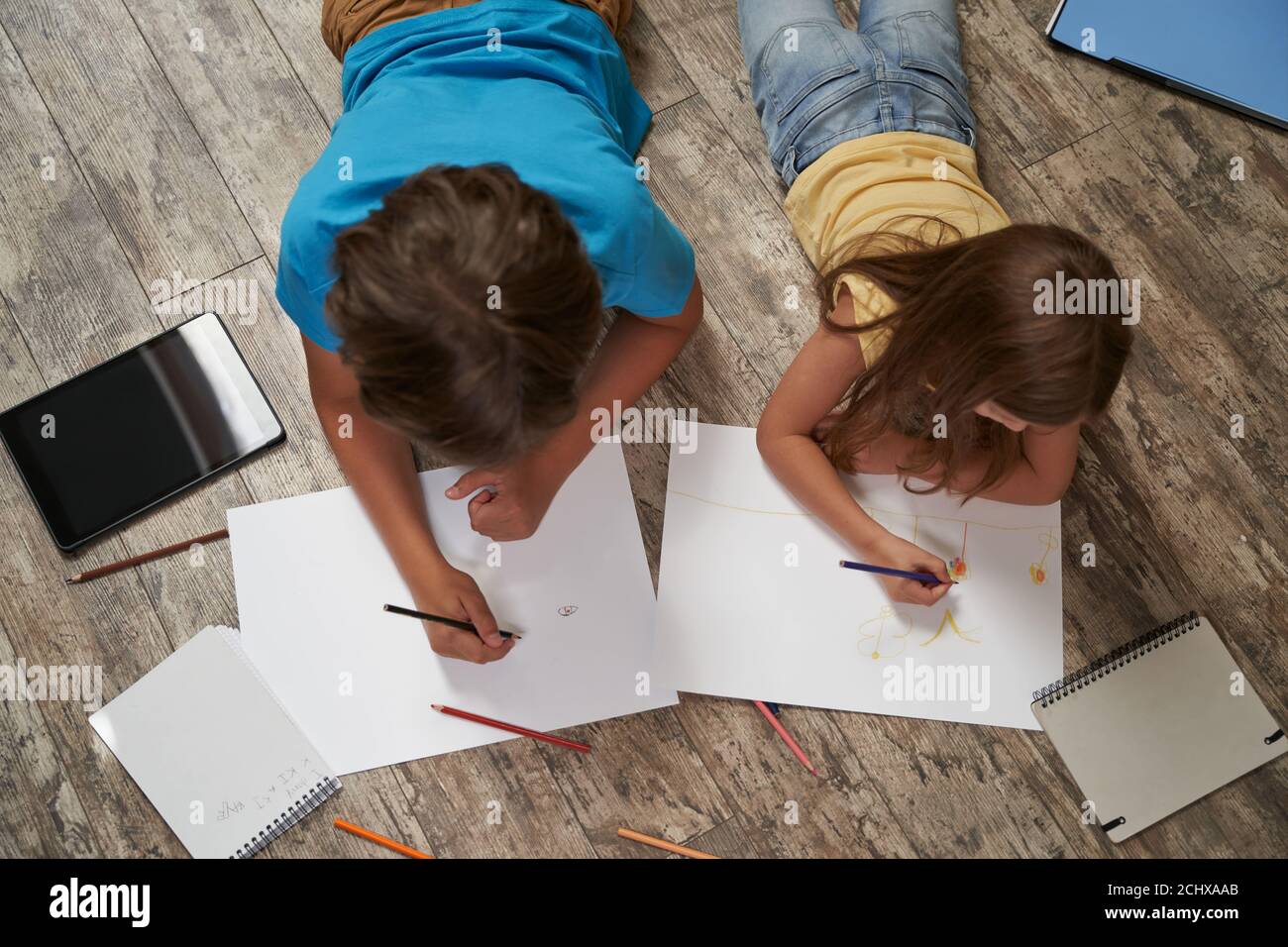 Fratelli che giocano insieme a casa. Vista dall'alto del ragazzino e della ragazza che giacciono sul pavimento di legno e disegnano su fogli bianchi di carta con colorato Foto Stock