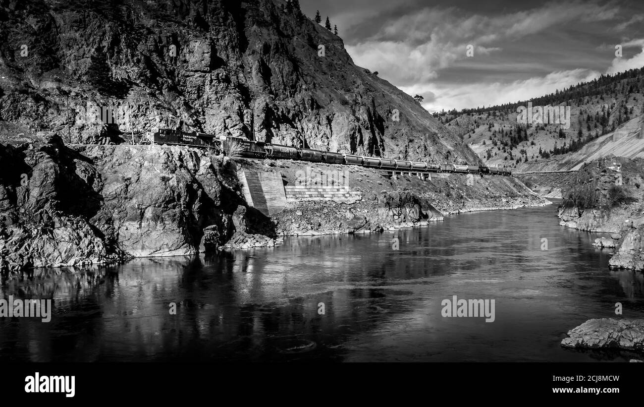 Foto in bianco e nero di un lungo treno cargo in viaggio Lungo le ripide scogliere del Thompson River Canyon Foto Stock