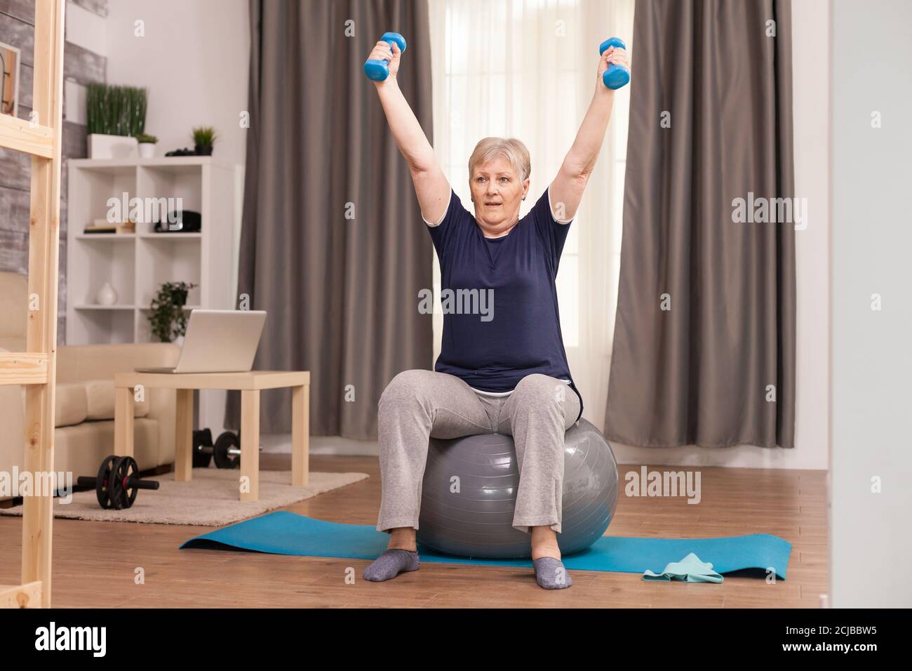 Nonna con stile di vita sano che pratica il fitness a casa. Anziano pensionato online internet esercizio di formazione a casa attività sportiva con manubri, banda di resistenza, palla svizzera all'età di anziano pensionamento Foto Stock