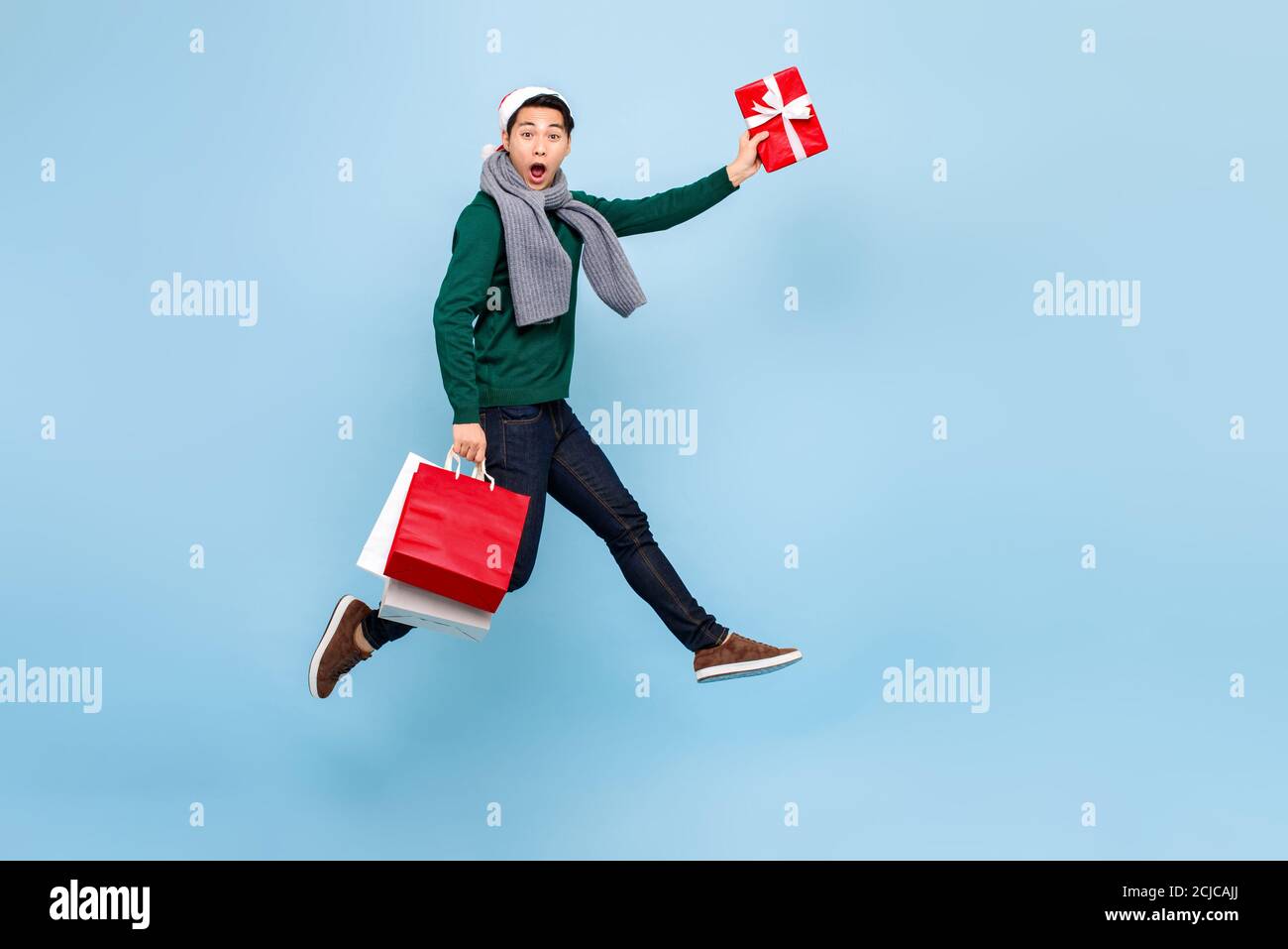 Ritratto a lunghezza intera di felice giovane bell'uomo asiatico dentro abbigliamento natalizio che salta mentre si tiene in mano borsa rossa e regalo scatola isolata in blu chiaro Foto Stock