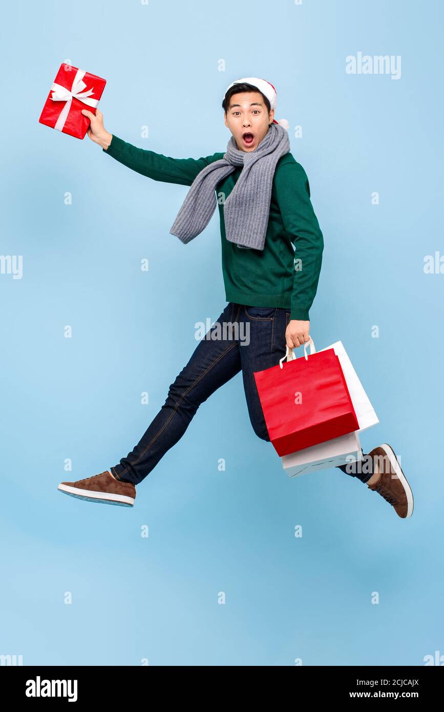 Ritratto a lunghezza intera di felice giovane bell'uomo asiatico dentro abbigliamento natalizio che salta mentre si tiene in mano borsa rossa e regalo scatola isolata in blu chiaro Foto Stock