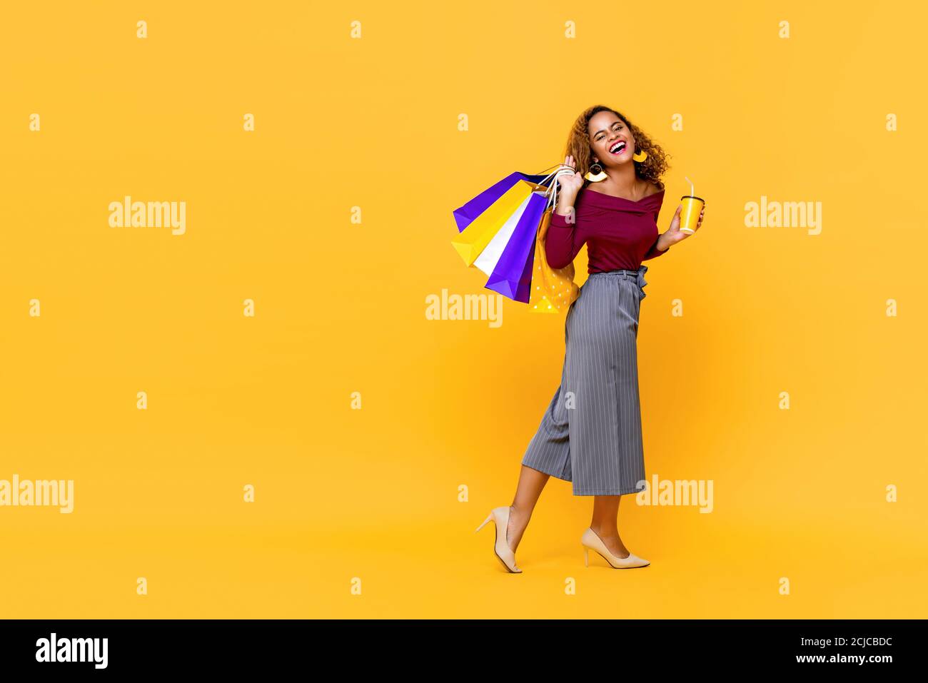 Ritratto di una giovane donna afroamericana sorridente che tiene borse per lo shopping e una tazza di bevande camminando in giallo studio isolato sfondo con cop Foto Stock