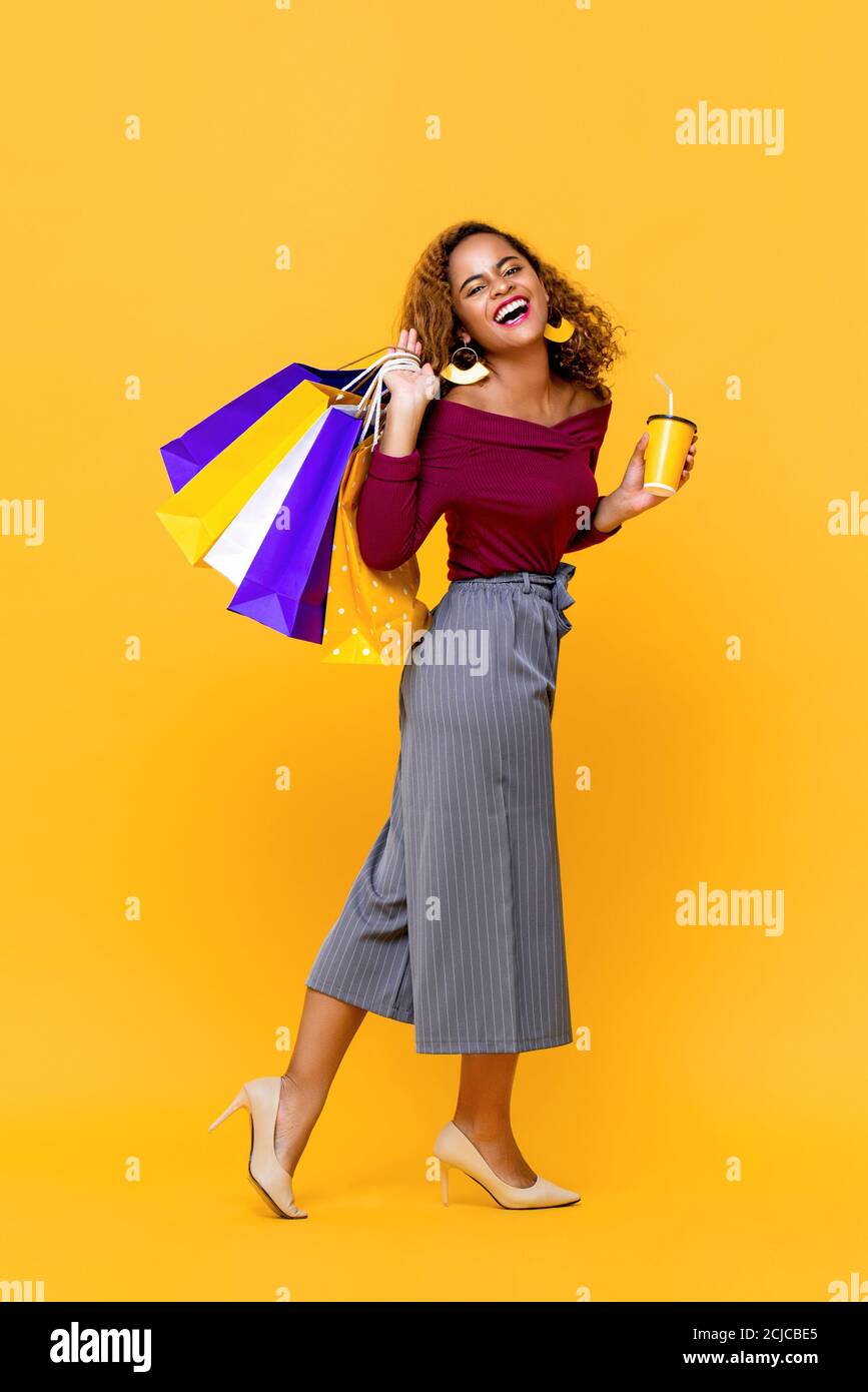 Ritratto di una giovane donna afroamericana sorridente che tiene borse per lo shopping e una tazza di bevande camminando in giallo studio isolato sfondo Foto Stock