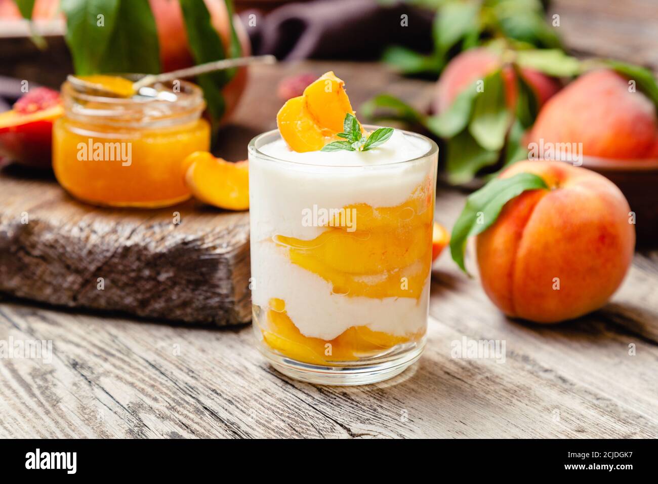 Dessert alla frutta di pesca in una tazza di vetro su un tavolo rustico in legno con frutta fresca di pesca, marmellata di pesche. Dessert fatto in casa con frutta. Macedonia di frutta con yogurt o. Foto Stock