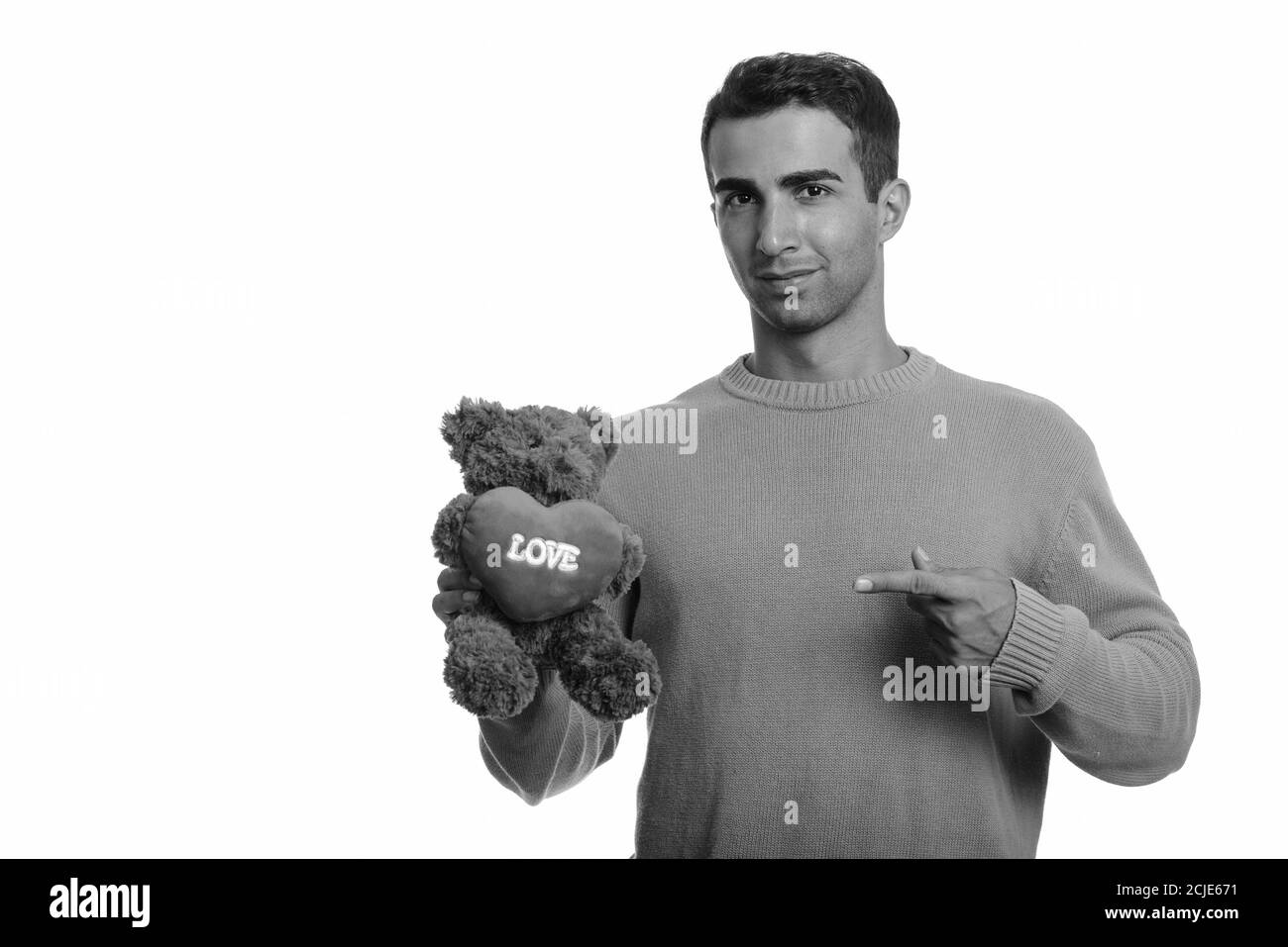 Giovane bell'uomo iraniano che tiene orsacchiotto con cuore e. Segno d'amore mentre punta il dito pronto per San Valentino Foto Stock