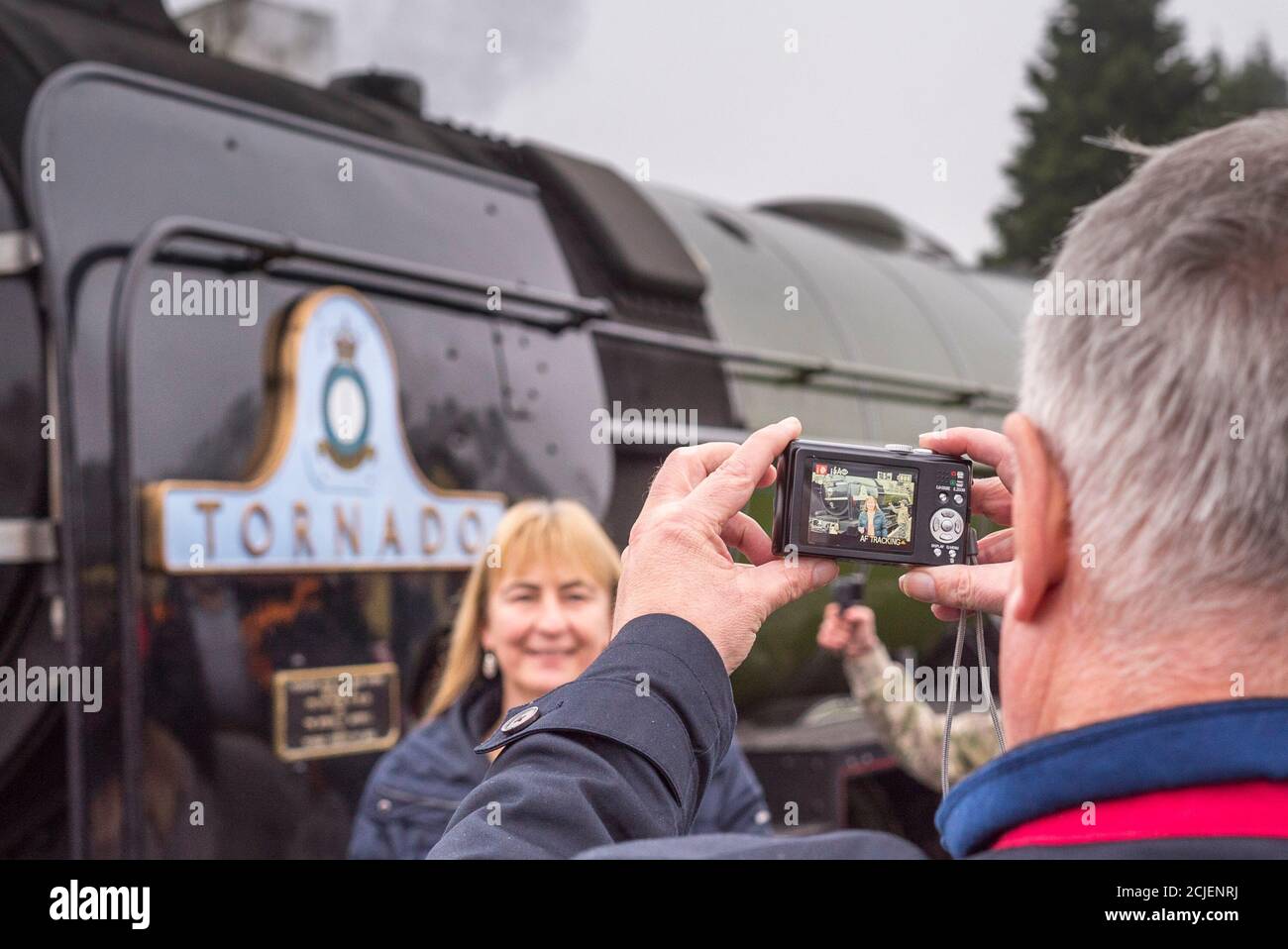 Uomo di mezza età che fotografa sua moglie, signora in piedi di fronte alla locomotiva a vapore britannica Tornado 60163. Foto Stock