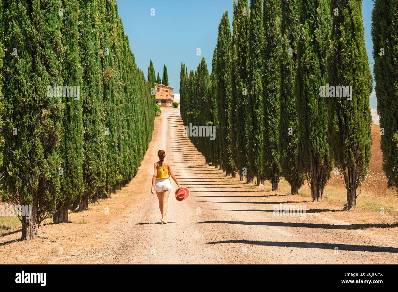 Vista posteriore di una donna che cammina lungo una strada sterrata fiancheggiata da cipressi toscani in una giornata di sole. Villa italiana all'orizzonte con sfondo blu. Foto Stock