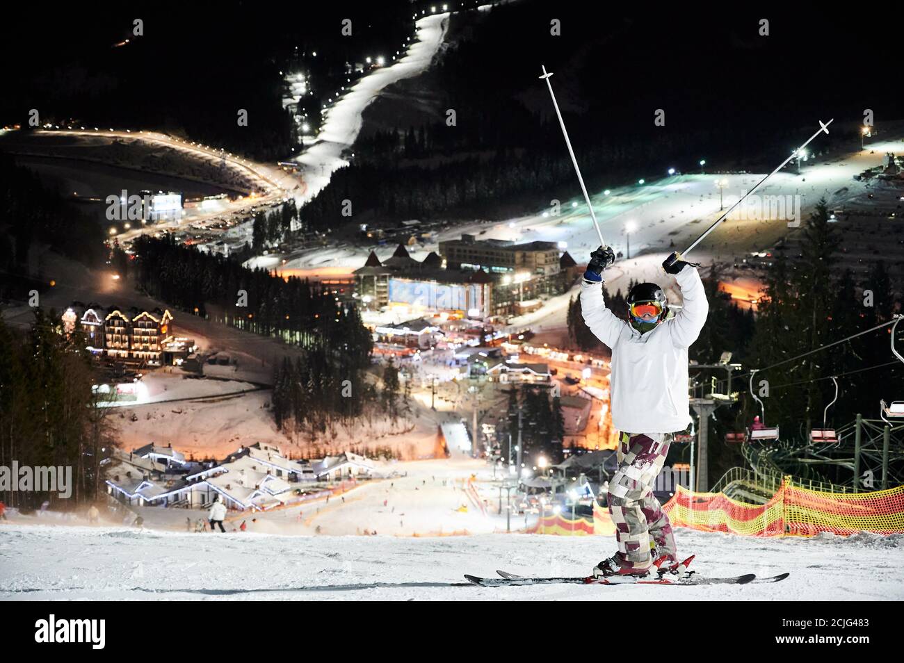 Lunghezza completa di sciatore maschile in giacca invernale e casco che solleva pali da sci e guardando la macchina fotografica. Giovane uomo in occhiali da sci in piedi su una pista innevata con bella città notturna sullo sfondo Foto Stock