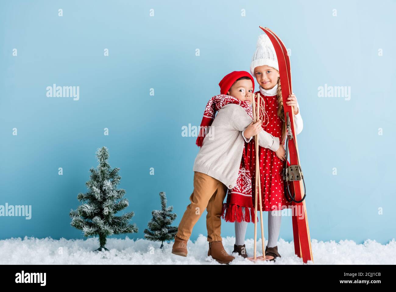 ragazzo in cappello e sciarpa hugging sorella con bastoni da sci e sci mentre si è in piedi su blu Foto Stock