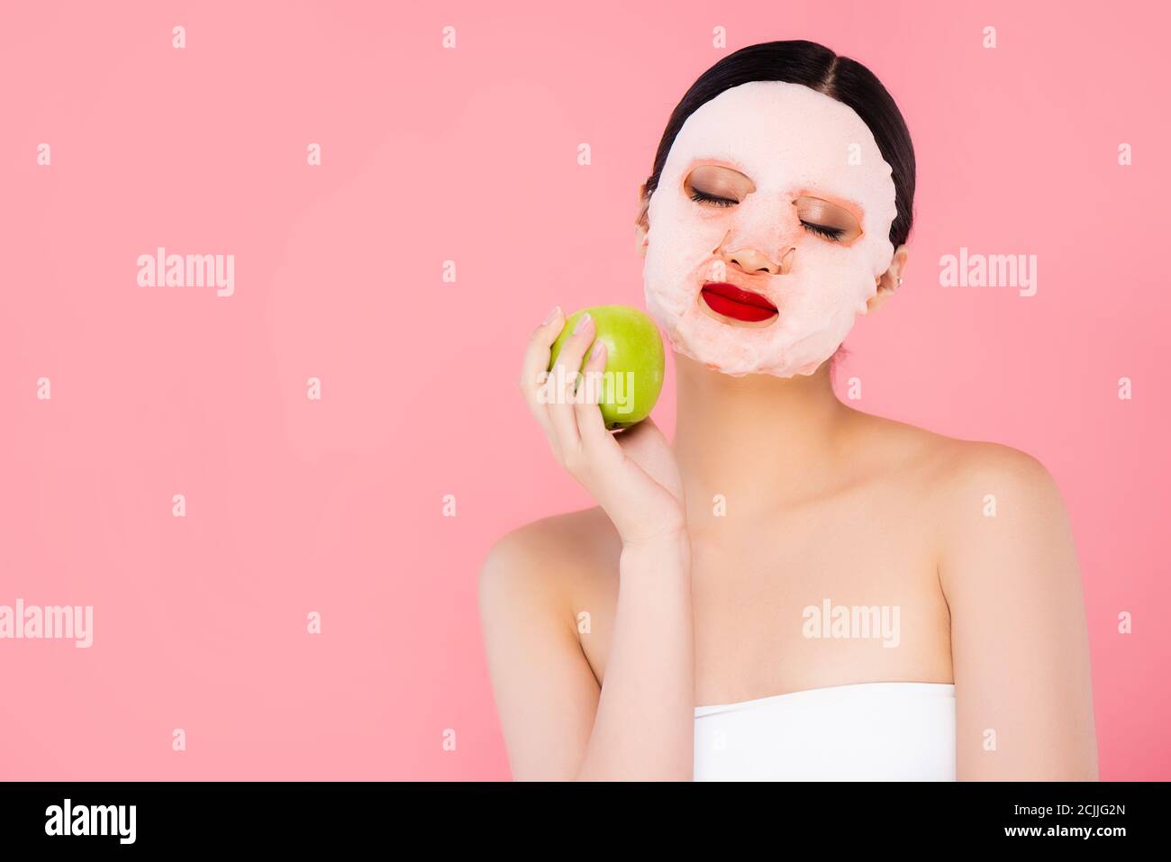 giovane donna asiatica in maschera di faccia che tiene la mela verde con occhi chiusi isolati su rosa Foto Stock