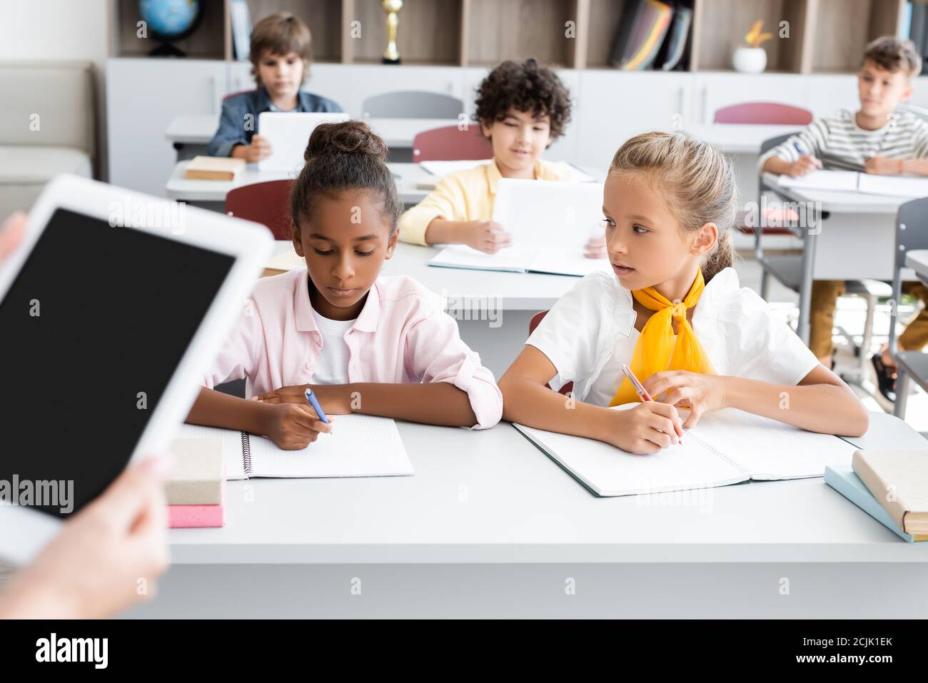 vista ritagliata del tablet digitale con supporto per insegnanti e schermo vuoto vicino agli allievi multiculturali che scrivono nei taccuini Foto Stock