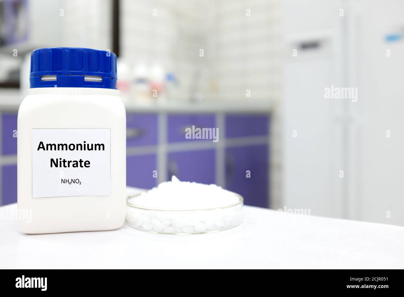 Fuoco selettivo di una bottiglia di nitrato di ammonio in una bottiglia di plastica accanto a una capsula di Petri con palline bianche all'interno di un fondo di laboratorio. Foto Stock