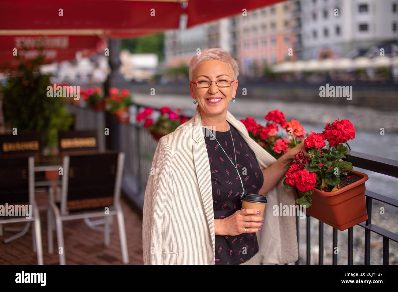 donna felice con fiori meravigliosi sul piatto sta posando alla macchina fotografica nel caffè di strada. case meravigliose e fiume sullo sfondo sfocato Foto Stock