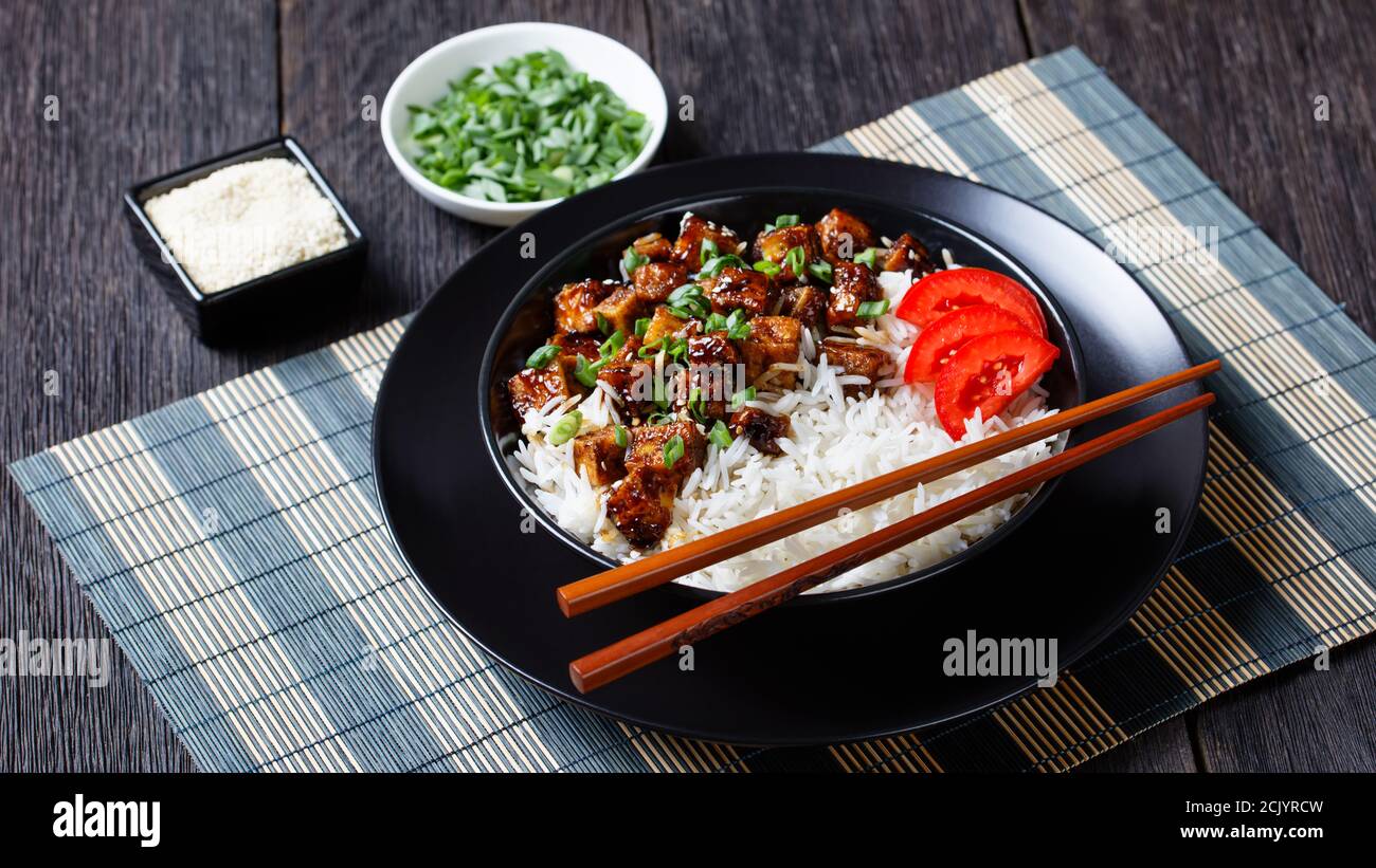 Ricetta a base di piante di tofu teriyaki con riso cosparso di scalione, semi di sesamo serviti con riso, pomodoro a fette, su una ciotola nera con bastoncini Foto Stock