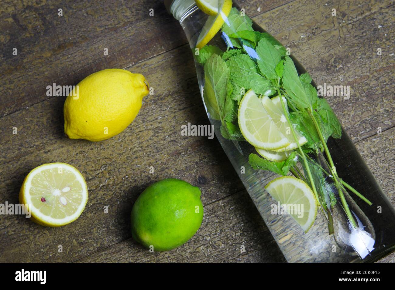 Vista su una bottiglia di vetro dell'acqua isolata aromatizzata con foglie di menta e agrumi gialli, frutti di lime verdi su un tavolo di legno (Focus su bottiglia) Foto Stock