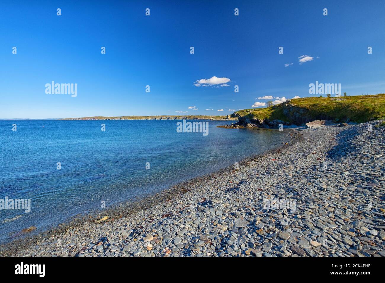 Cielo blu luminoso con alcune nuvole bianche su una spiaggia di ciottoli con alcune alghe innaffate sulla riva. C'è una massa di terra o punto alla fine della spiaggia. Foto Stock
