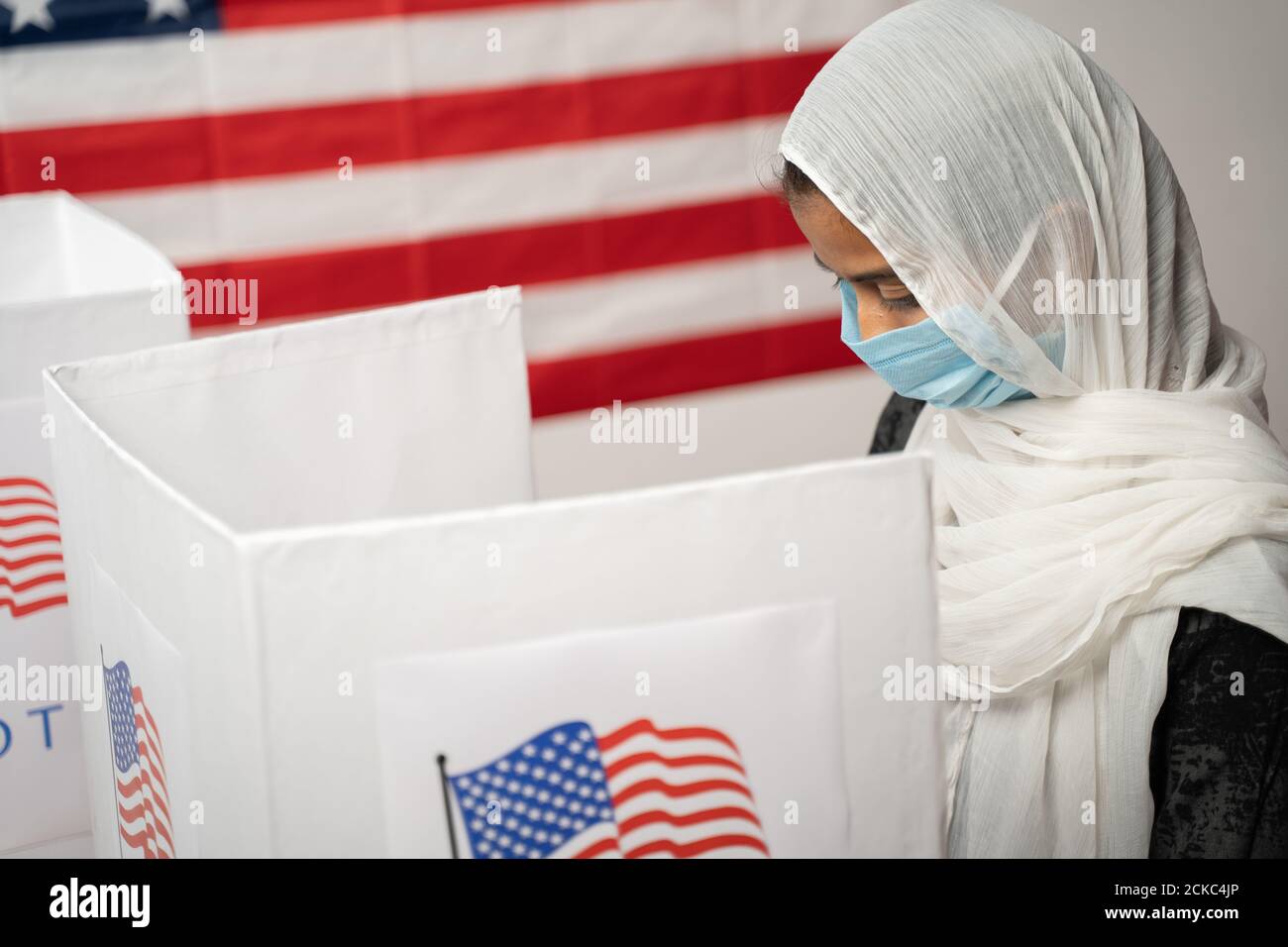 Ragazza con Hijab o copertura testa e maschera indossata occupato al banco di polling con la bandiera degli Stati Uniti come sfondo - concetto di elezione degli Stati Uniti. Foto Stock
