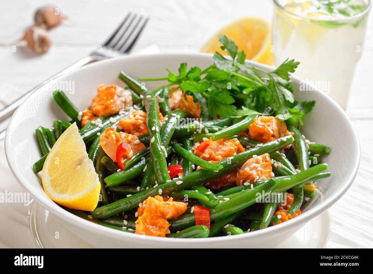 Salmone al curry con fagioli verdi, peperoni rossi cosparsi di semi di sesamo serviti su un piatto bianco con prezzemolo, zeppa di limone, su un bac di legno bianco Foto Stock