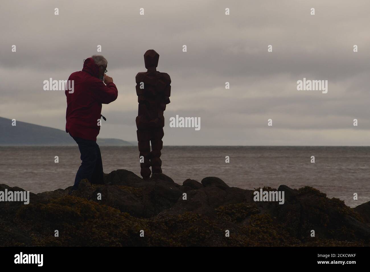 La scultura, IL GRIP di Anthony Gormley e un uomo 60+ in piedi su rocce a Saddell Bay, Kintyre, Argyll, Scozia, guardando insieme Kilbrannan Sound Foto Stock