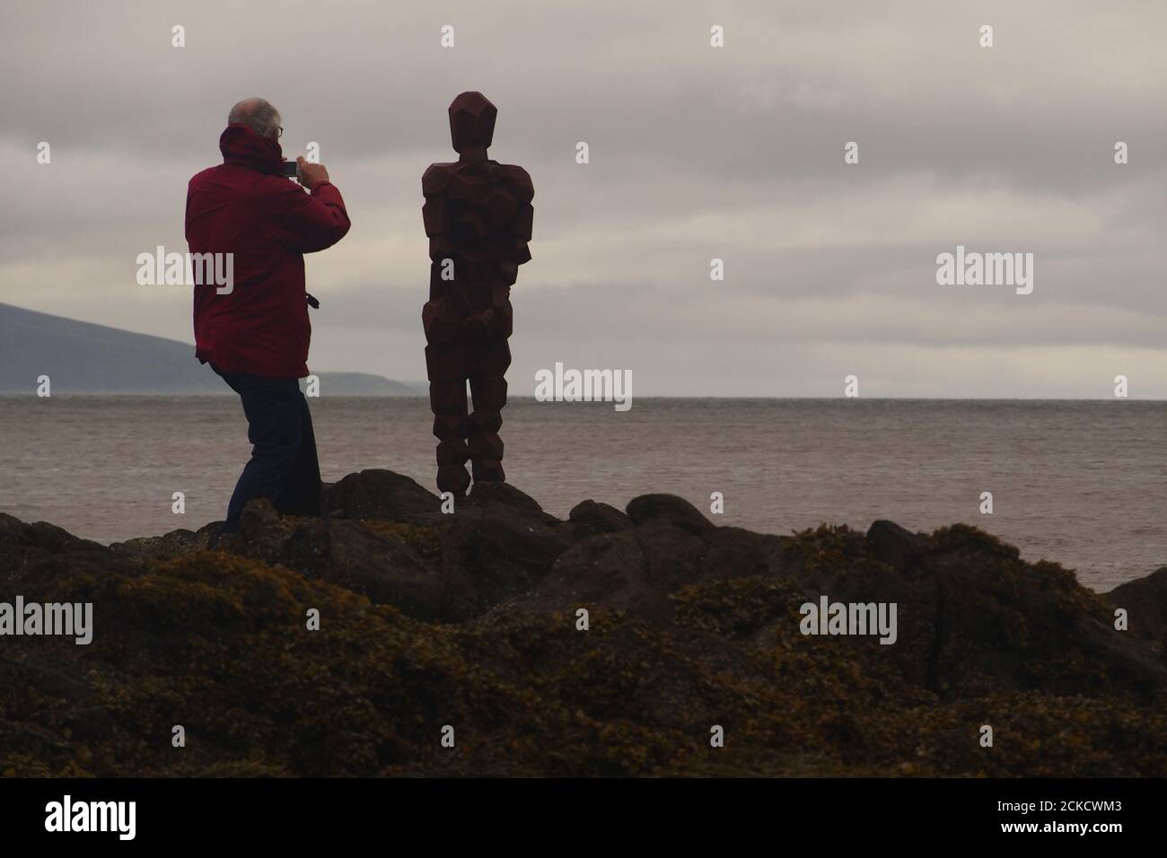 La scultura, IL GRIP di Anthony Gormley e un uomo 60+ in piedi su rocce a Saddell Bay, Kintyre, Argyll, Scozia, guardando insieme Kilbrannan Sound Foto Stock