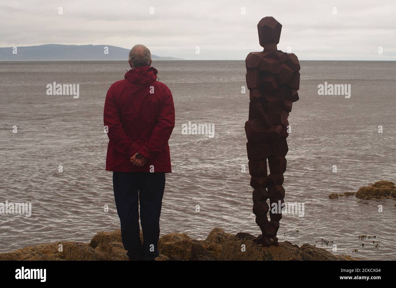 La scultura, IL GRIP di Anthony Gormley e un uomo 60+ in piedi su rocce a Saddell Bay, Kintyre, Argyll, Scozia, guardando insieme Kilbrannan Sound Foto Stock