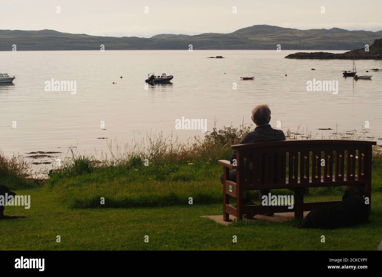Un uomo anziano 60+ seduto su una panchina che guarda verso il mare a Carsaig Bay, Tayvallich, Argyll, Scozia, in attesa dei cieli serali Foto Stock