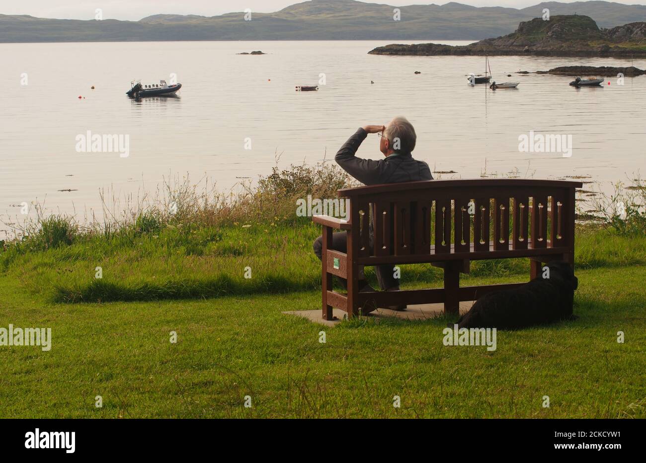 Un uomo anziano 60+ seduto su una panchina che guarda verso il mare a Carsaig Bay, Tayvallich, Argyll, Scozia, in attesa dei cieli serali Foto Stock