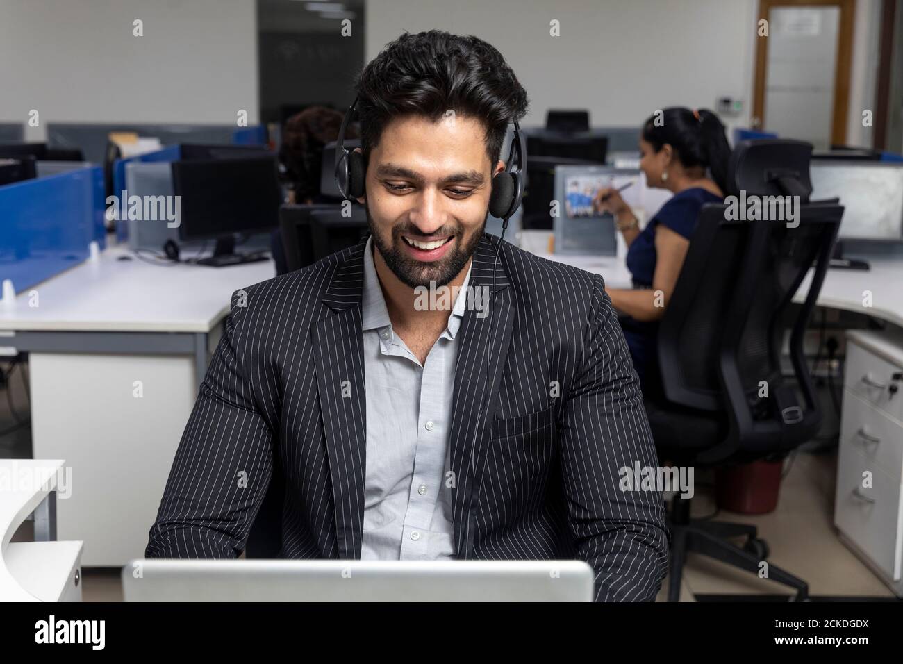 Ritratto di un giovane uomo d'affari indiano che indossa le cuffie, seduto sulla sua postazione di lavoro sullo sfondo dell'ufficio, ambiente aziendale, call center. Foto Stock