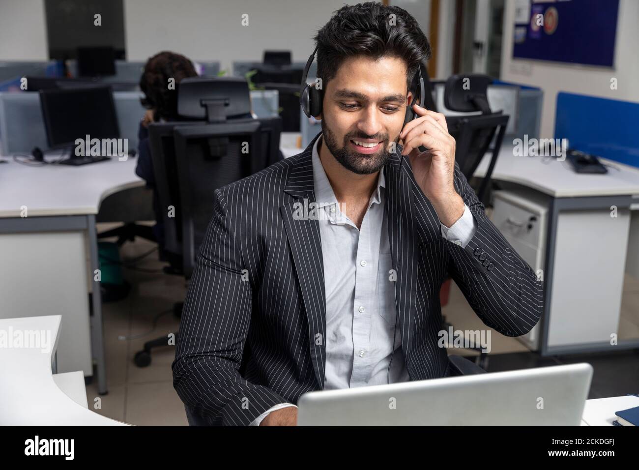 Ritratto di un giovane uomo d'affari indiano che indossa le cuffie, seduto sulla sua postazione di lavoro sullo sfondo dell'ufficio, ambiente aziendale, call center. Foto Stock