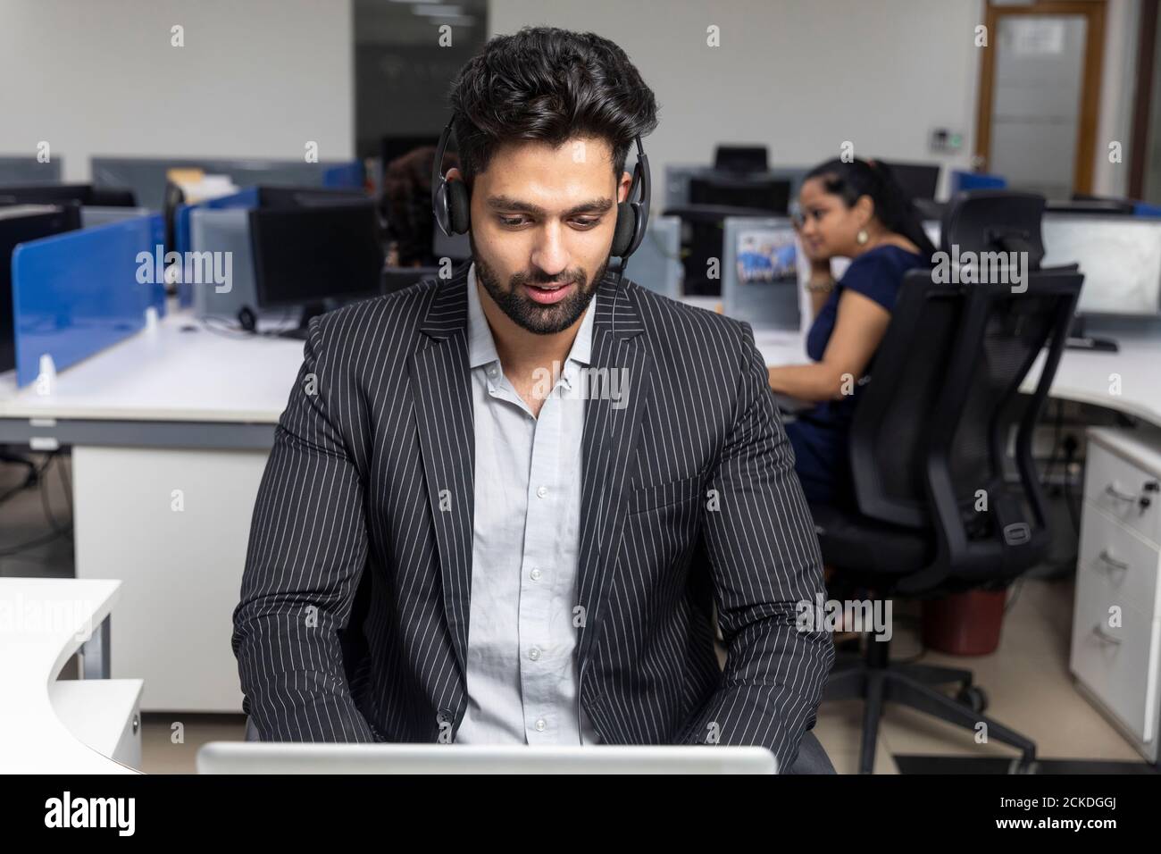 Ritratto di un giovane uomo d'affari indiano che indossa le cuffie, seduto sulla sua postazione di lavoro sullo sfondo dell'ufficio, ambiente aziendale, call center. Foto Stock