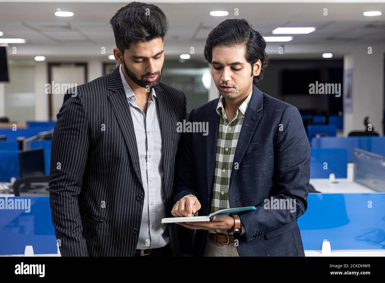 Ritratto di due giovani bei imprenditori indiani in piedi contro lo sfondo dell'ufficio, discutere di progetto, guardando al diario, ambiente aziendale. Foto Stock