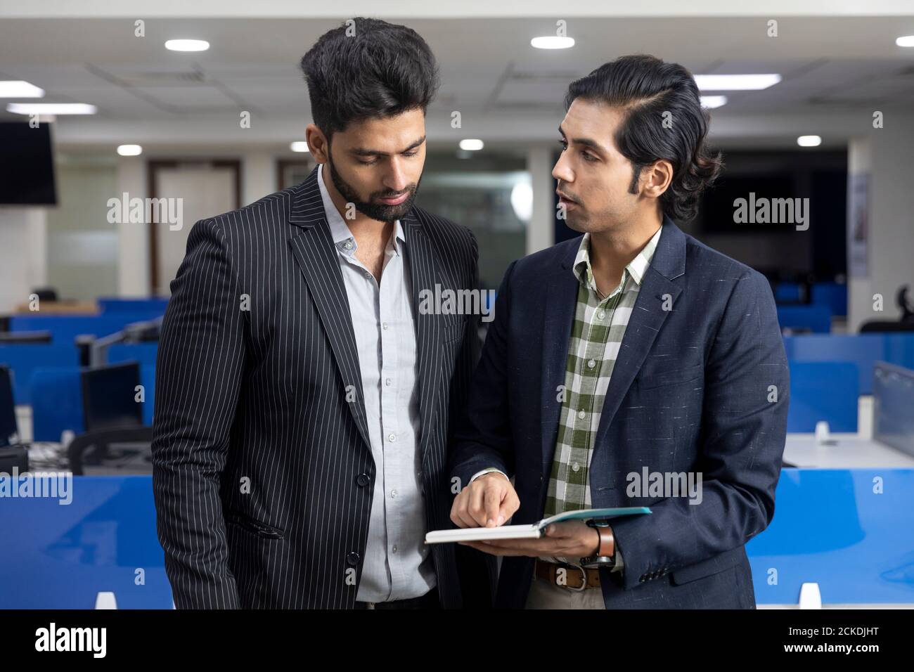 Ritratto di due giovani bei imprenditori indiani in piedi contro lo sfondo dell'ufficio, discutere di progetto, guardando al diario, ambiente aziendale. Foto Stock