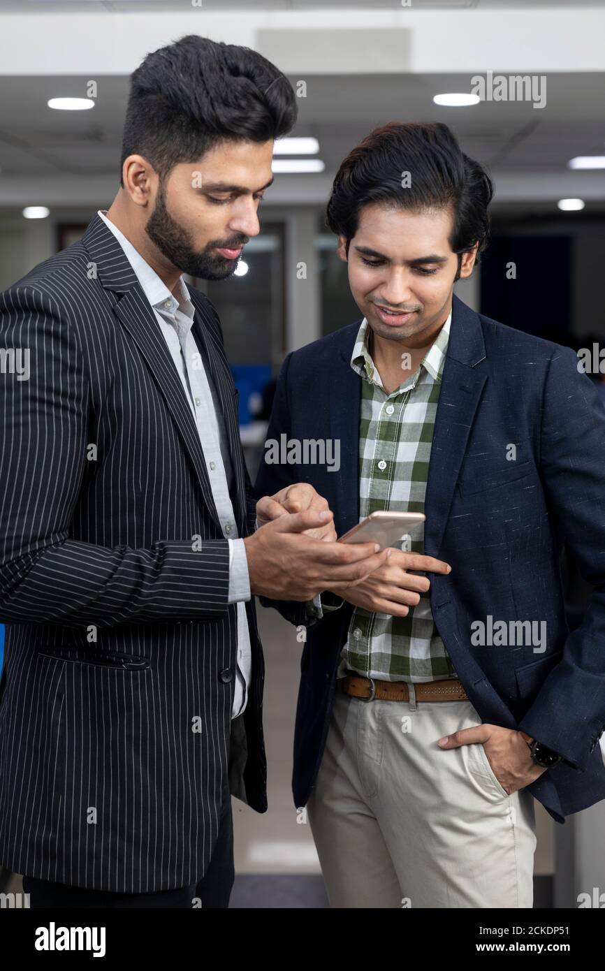 Ritratto di due giovani bei uomini d'affari indiani che guardano al telefono, discutono progetto, ambiente aziendale. Foto Stock