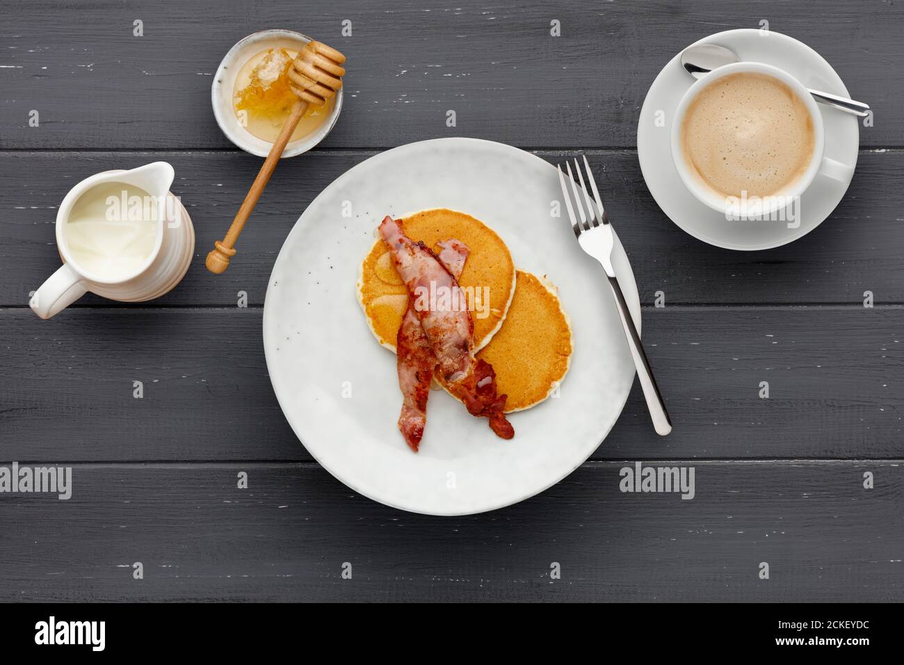 Un piatto di pancetta e frittelle, condito con miele, una tazza di caffè e una brocca di panna su uno sfondo di legno scuro Foto Stock