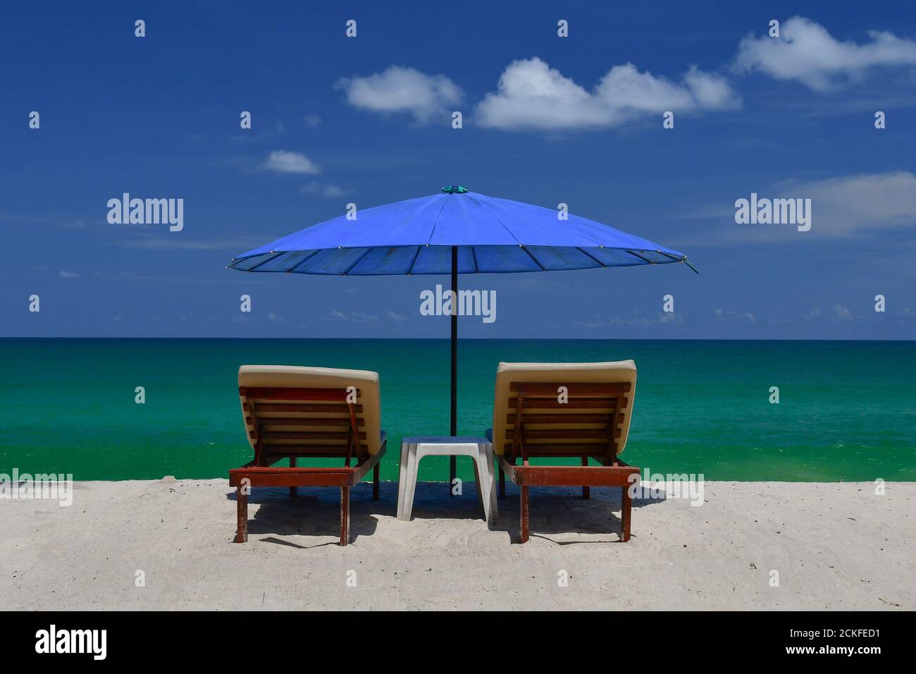 Vista posteriore di un paio di sedie a sdraio in legno a doghe su una spiaggia tropicale di sabbia bianca, sotto un ombrellone blu. Foto Stock