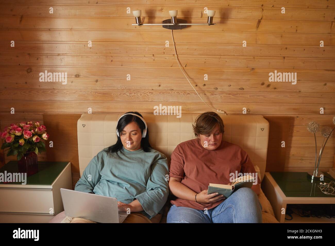 Due giovani donne in sovrappeso che si rilassano a letto con computer portatile e. prenota in camera da letto Foto Stock