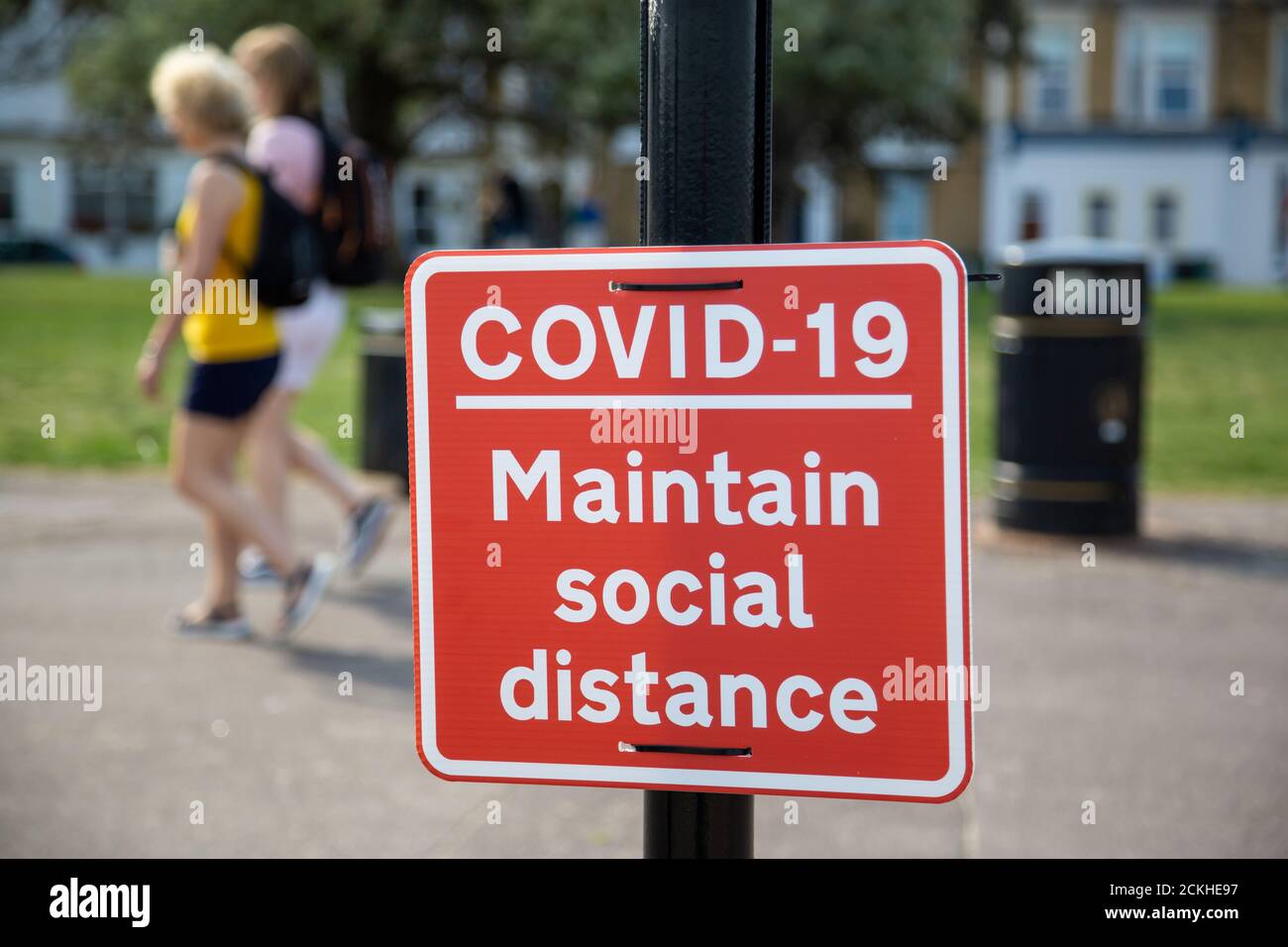 Persone che camminano oltre un cartello su un lampione che legge Covid-19 mantenere la distanza sociale Foto Stock