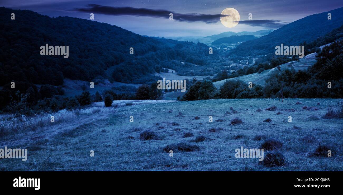 strada di campagna attraverso campo rurale di notte. paesaggio suburbano estivo in montagna in piena luce luna. villaggio nella valle lontana. giorno nuvoloso Foto Stock