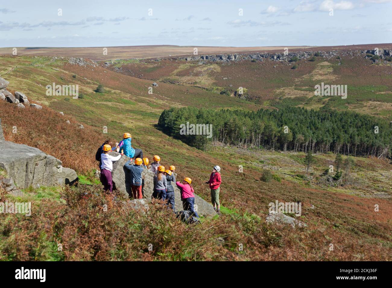 I bambini in caschi di sicurezza arrampicano sotto istruzioni su massi in pietra con vista panoramica sulla Burbage Valley, Sheffield Foto Stock