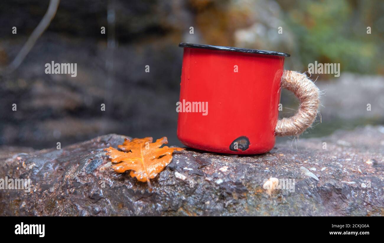 Tazza di smalto rosso acquerello rimanere sulla roccia vicino alla sorgente d'acqua. Stile vintage retrò. Tazza carina in metallo. Foto Stock