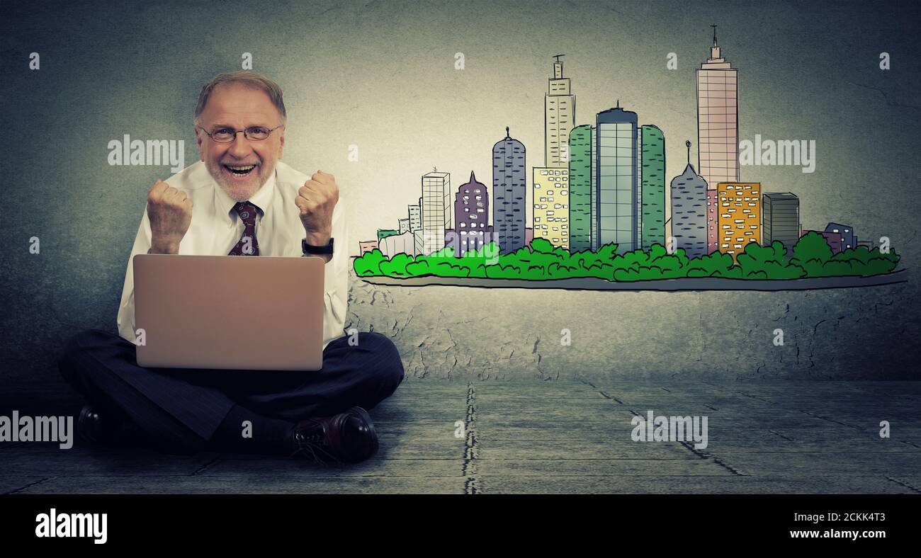 Eccitato anziano uomo seduto con notebook tenendo i pugni fino felice con real estate contro uno sfondo grigio Foto Stock