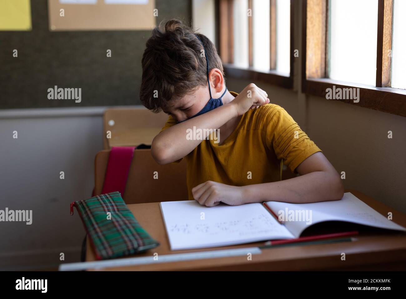 Ragazzo che indossa la maschera che starnutisce mentre si siede sulla sua scrivania a scuola Foto Stock