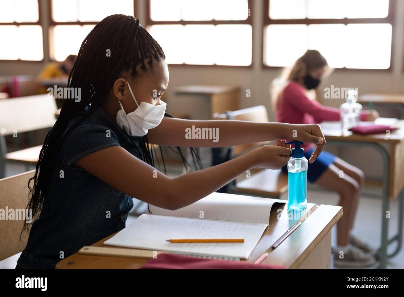 Ragazza che indossa maschera viso igienizzante le mani mentre si siede la sua scrivania a scuola Foto Stock