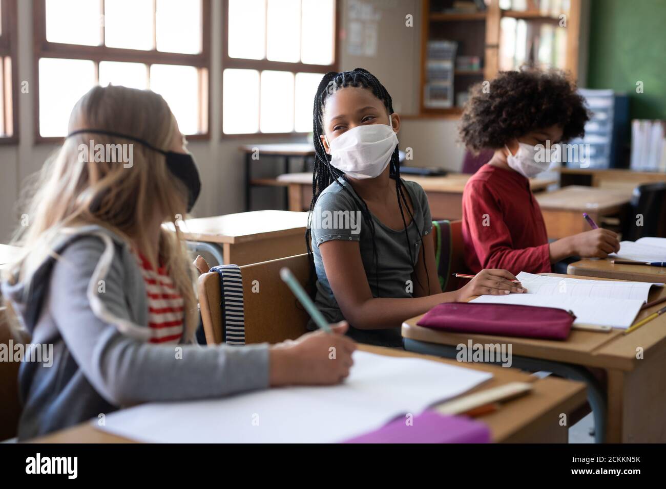 Due ragazze che indossano maschere di faccia che parlano l'una con l'altra mentre seduti sulla scrivania a scuola Foto Stock
