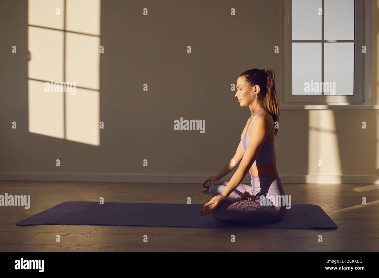 Yoga donna in classe yoga pratica yoga seduto sul pavimento in posizione lotus in una camera con finestre soleggiate. Foto Stock