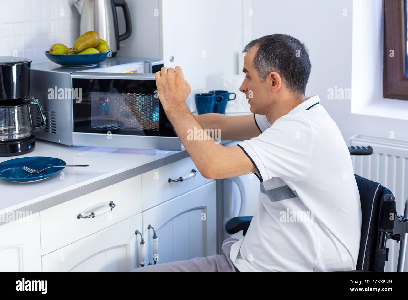 Giovane uomo in Whellchair che usa forno a microonde per cuocere in cucina. Mettere a fuoco sulla sua mano. Foto Stock