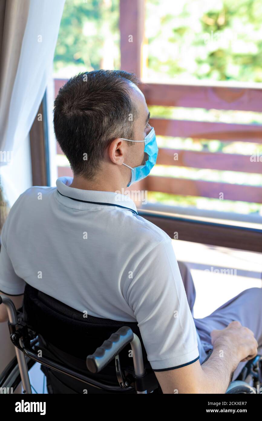 Solo giovane uomo che indossa una maschera seduta su una sedia a rotelle che guarda fuori dalla finestra. Mettere a fuoco sulla testa. Foto Stock Solo giovane uomo che indossa una maschera seduta su una sedia a rotelle che guarda fuori dalla finestra. Mettere a fuoco sulla testa. Foto Stock