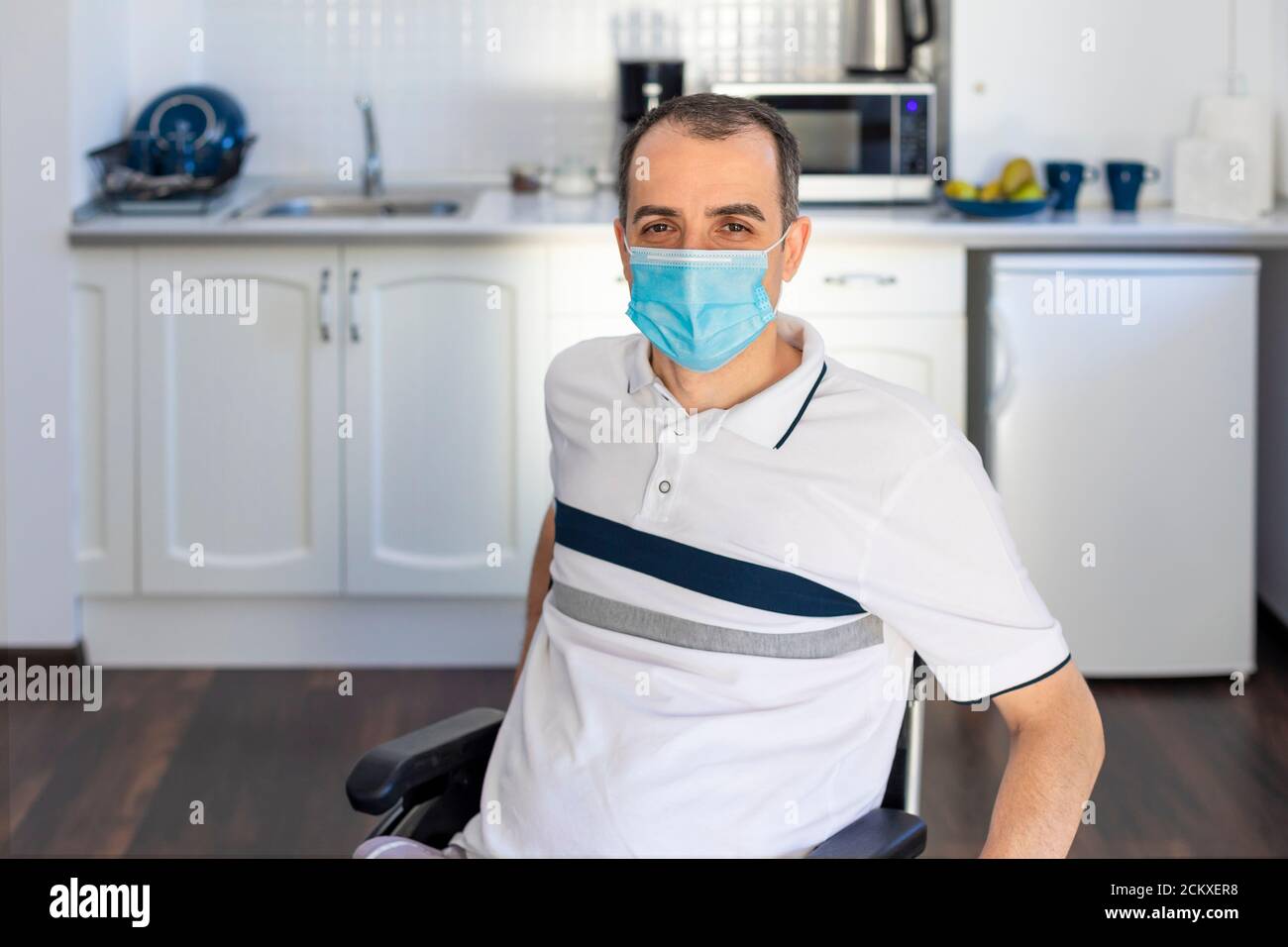 Sorridente giovane uomo portatore seduto su sedia a rotelle in cucina. Giovane uomo che indossa la maschera seduta davanti alla cucina. Mettere a fuoco sul suo volto. Foto Stock Sorridente giovane uomo portatore seduto su sedia a rotelle in cucina. Giovane uomo che indossa la maschera seduta davanti alla cucina. Mettere a fuoco sul suo volto. Foto Stock